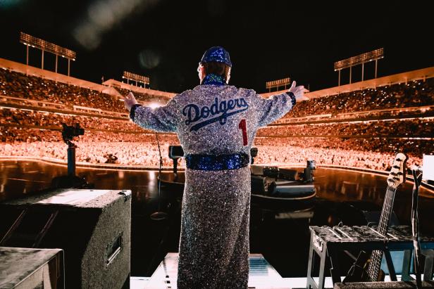 Elton John von hinten fotografiert. Er steht in glitzerndem Mantel auf der Bühne. Vor ihm das volle Dodgers-Stadion in Los Angeles.