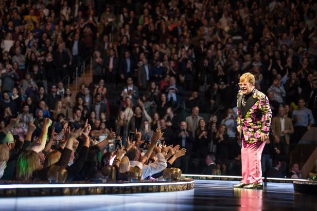 Elton John im Blumensakko auf der Bühne. Er steht nahe bei den Fans, die ihm zujubeln.