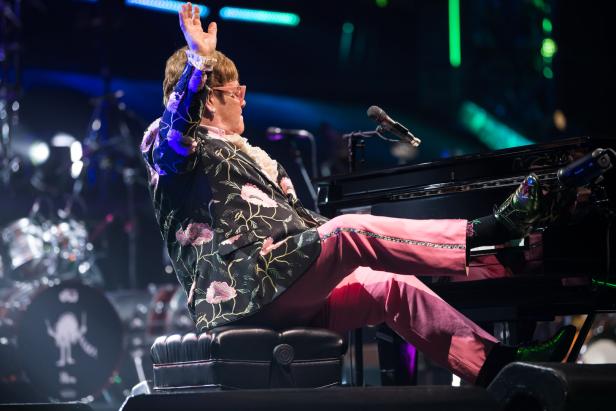 Elton John sitzt am Klavier, er lehnt sich leicht zurück und schmeißt seinen rechten Fuß auf die Tastatur.