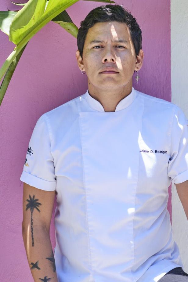 Ein Mann, Jaime D. Rodriguez, in Kochuniform vor einer rosa Wand mit Pflanzen.