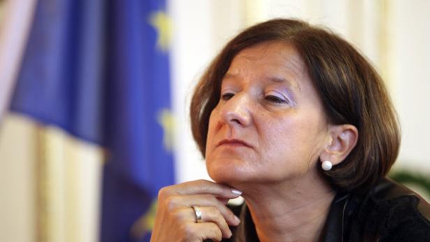 Eine Frau mit kurzem braunem Haar und Ohrringen vor einer EU-Flagge.