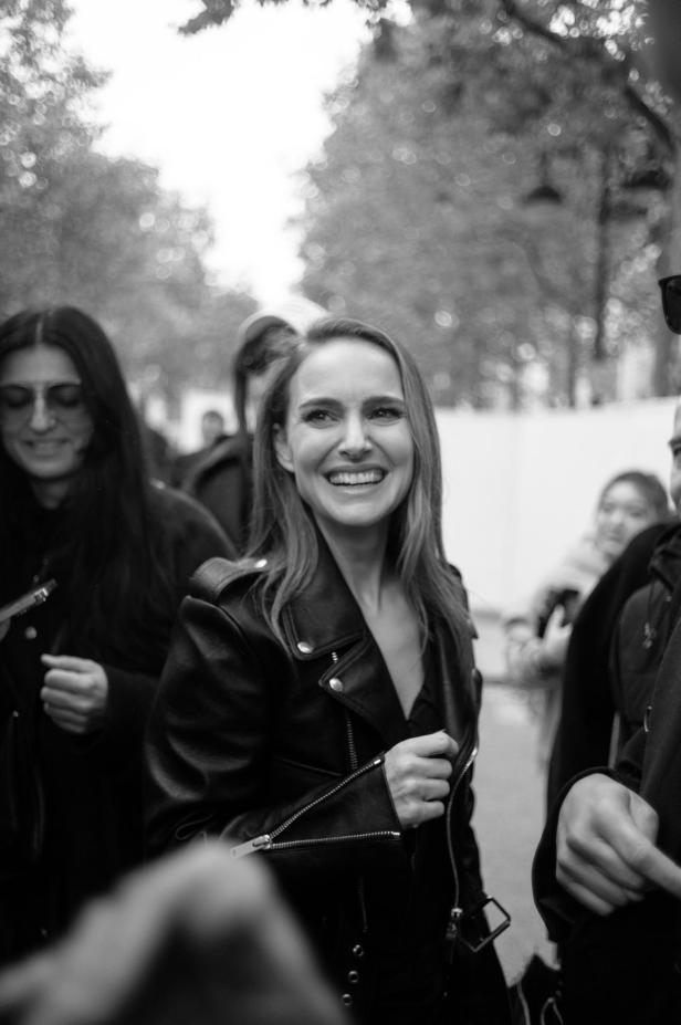 Natalie Portman lächelt in einer schwarzen Lederjacke, umgeben von anderen Menschen.