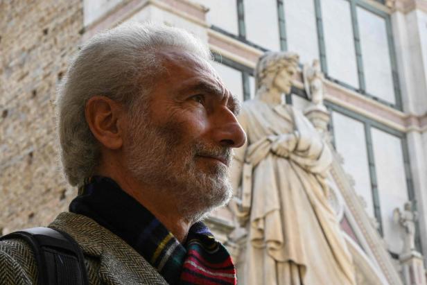 Ein Mann mit grauem Haar steht vor einer Statue, möglicherweise eine Darstellung von Dante.