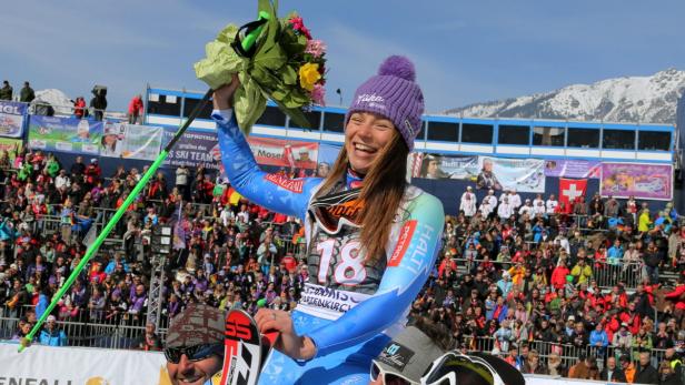 Eine strahlende Skirennläuferin mit Blumenstrauß auf dem Siegerpodest vor jubelnden Zuschauern.