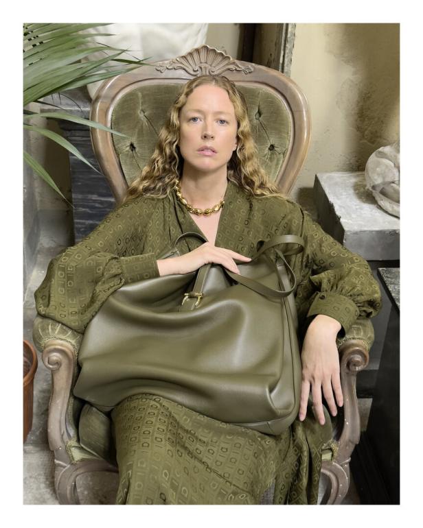 Eine Frau in einem grünen Kleid sitzt mit einer großen, olivgrünen Handtasche auf einem verzierten Stuhl.