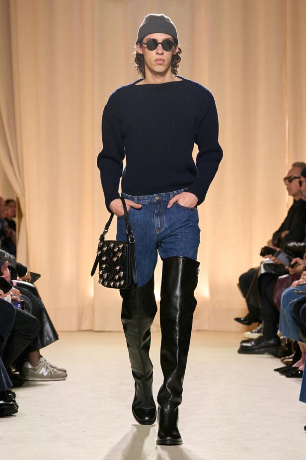 Ein Mann mit Mütze, Sonnenbrille, Pullover, Jeans und hohen Stiefeln läuft auf einem Laufsteg.