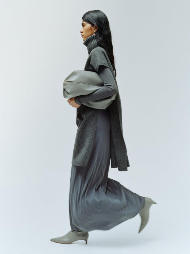 Eine Frau in einem grauen Ensemble mit Rollkragenpullover, Weste, Rock und Stiefeletten hält eine übergroße Tasche.