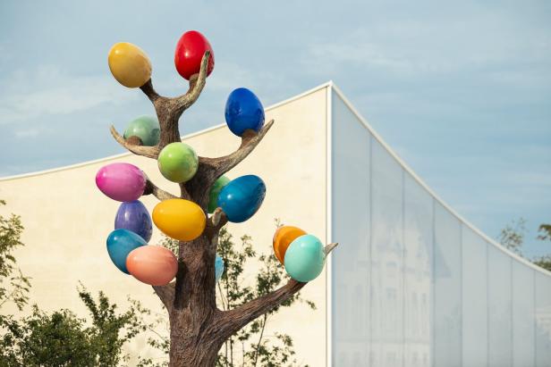 Ein Baum mit vielen bunten Perlen, die an Weintrauben erinnern sollen. Er steht vor einem neuen Gebäude, das sich wellt.