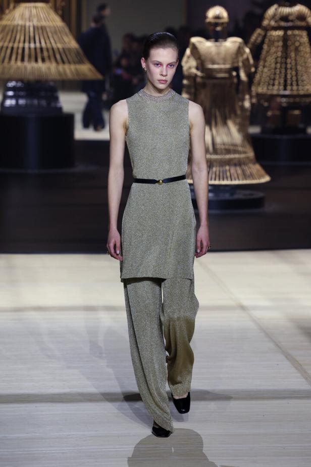 Ein Model präsentiert ein goldenes Hosen- und Top-Ensemble mit schwarzem Gürtel auf dem Laufsteg.