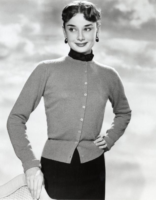Audrey Hepburn posiert in einem grauen Cardigan und einem schwarzen Rock vor einem bewölkten Himmel.