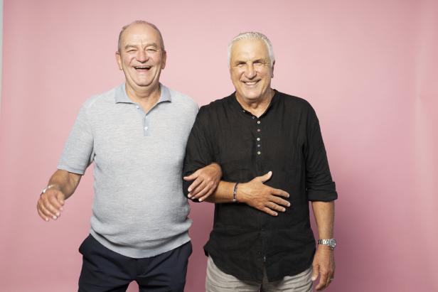 Zwei ältere Männer stehen Arm in Arm vor einem rosa Hintergrund und lachen.