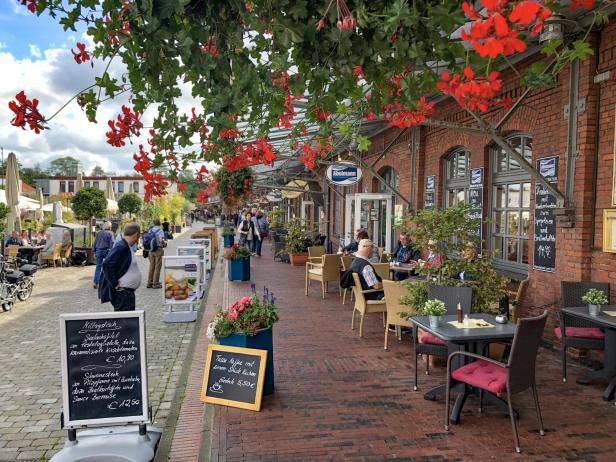 Mehrere Restaurants in Backsteinhäusern nebeneinander; mit Speisekarte-Schildern und roten, hängenden Blumen.