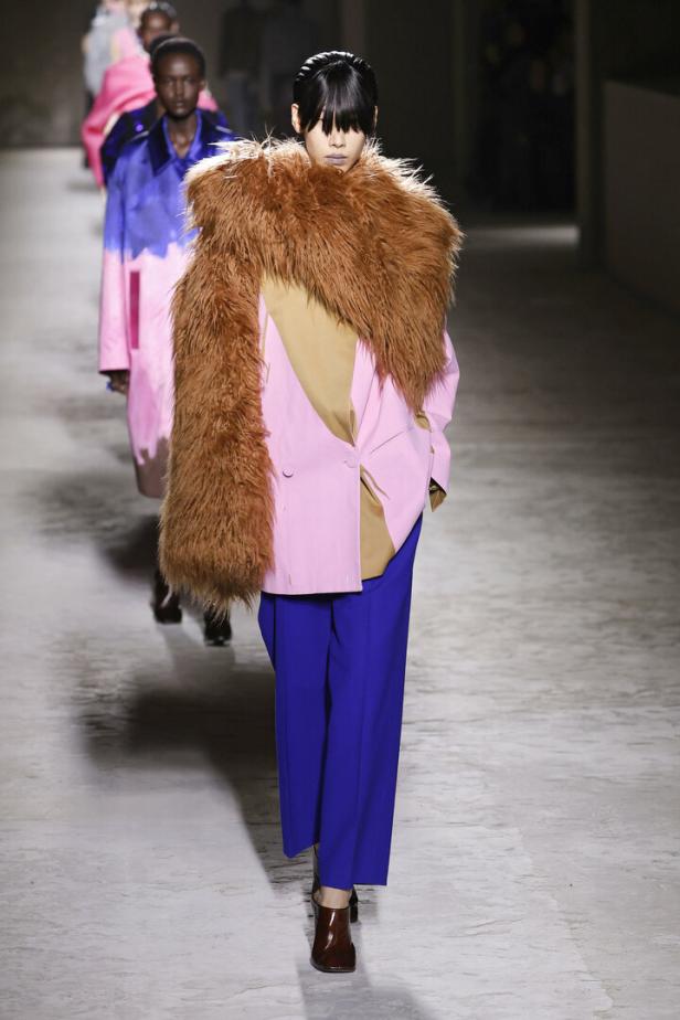 Eine Frau mit Pony trägt einen rosa Blazer mit braunem Fellkragen und eine blaue Hose auf dem Laufsteg.