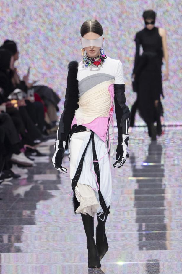 Ein Model mit futuristischer Brille präsentiert ein avantgardistisches Outfit auf dem Laufsteg.