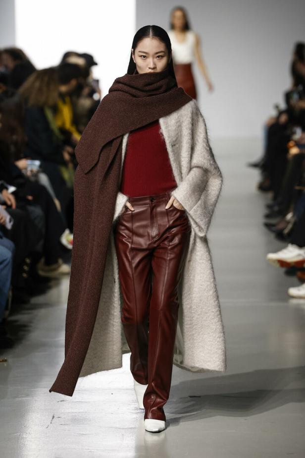 Eine Frau mit einem übergroßen Schal, einem roten Pullover, einer Lederhose und einem Mantel auf dem Laufsteg.