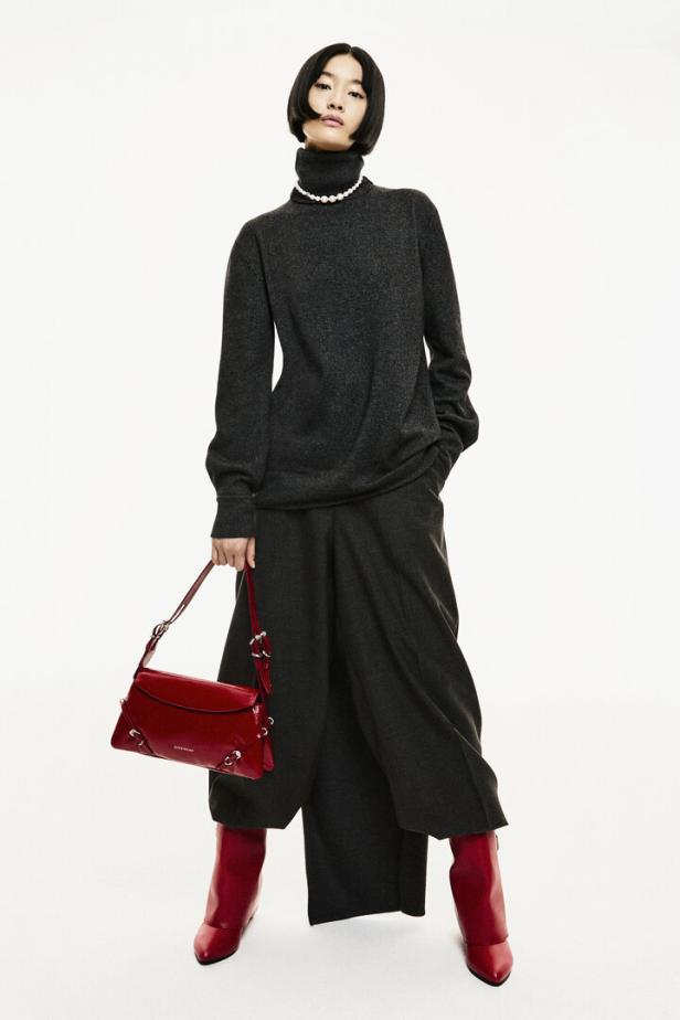 Eine Frau mit Perlenkette trägt einen grauen Rollkragenpullover, weite Hosen, rote Stiefel und eine rote Handtasche.