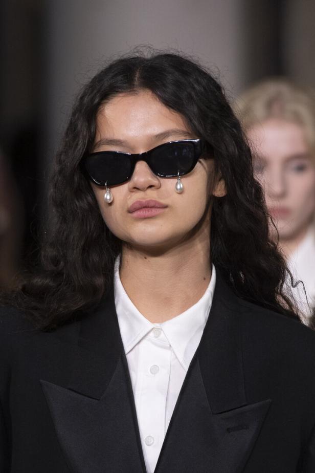 Eine Frau mit dunklen, gewellten Haaren trägt eine schwarze Brille mit Perlenanhängern.