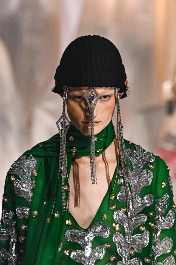 Ein Model mit einer schwarzen Mütze und einem verzierten, grünen Kleid mit silbernen Details.