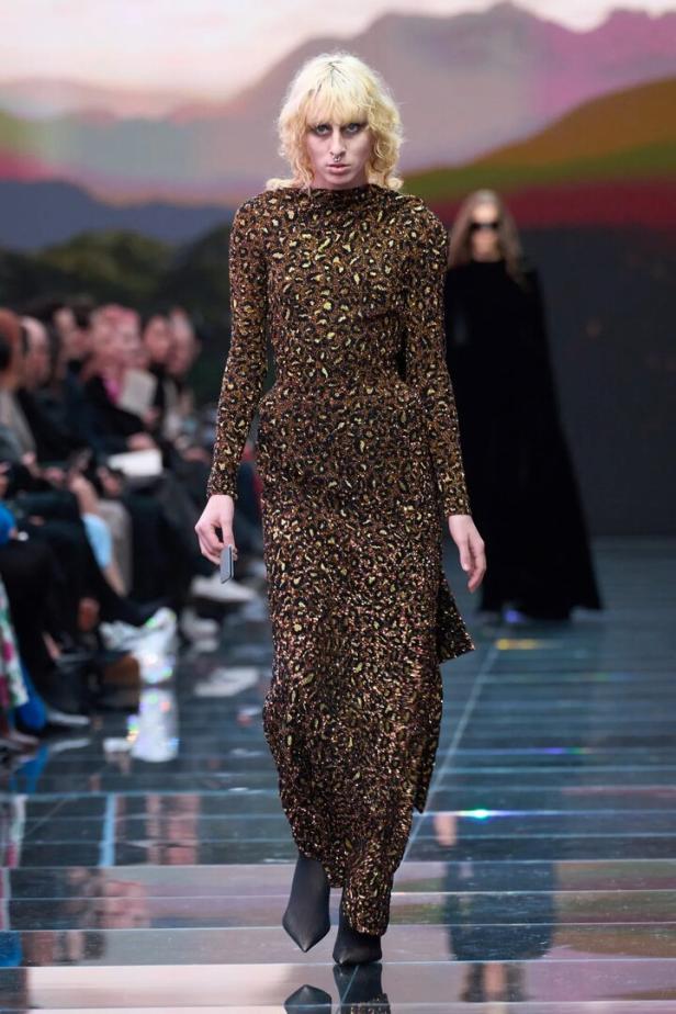 Ein Model mit blonden Haaren präsentiert ein langes, gemustertes Kleid auf einem Laufsteg.