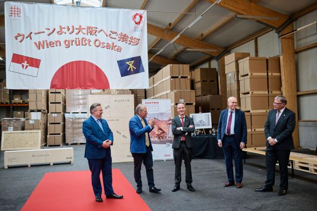 Fünf Männer stehen vor einem Banner „Wien grüßt Osaka“ in einer Lagerhalle.