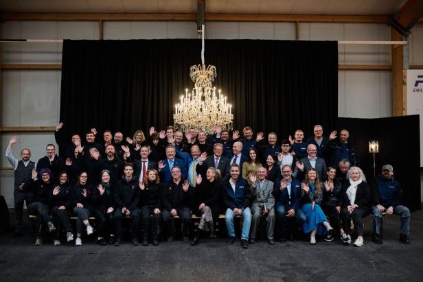Eine große Gruppe von Menschen winkt vor einem schwarzen Vorhang und einem Kronleuchter.