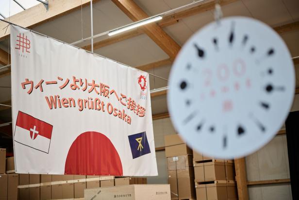 Ein Banner mit der Aufschrift „Wien grüßt Osaka“ und den Flaggen von Wien und Osaka hängt in einer Lagerhalle.