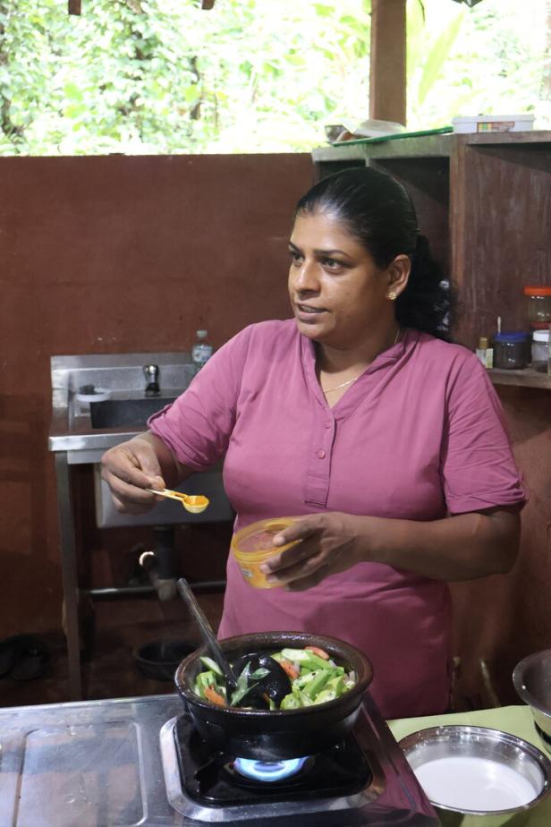 Eine Frau aus Sri Lanka kocht Gemüsecurry mit frischen Okraschoten und Chili über eine Gasherd und zeigt auf das Currypulver