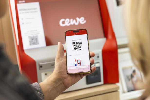 Auf einem Handy ist ein QR Code zu sehen. Hintendran sieht man einen der CEWE-Sofortfoto-Automaten.