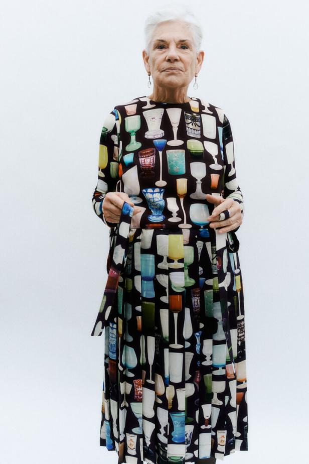 Eine ältere Frau mit grauen Haaren trägt ein Kleid mit einem Muster aus verschiedenen Gläsern.