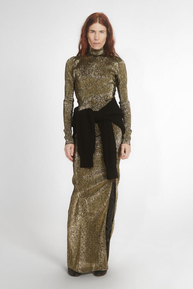 Eine Frau mit roten Haaren trägt ein goldenes, glitzerndes Kleid mit Rollkragen und einen schwarzen Pullover um die Hüften.