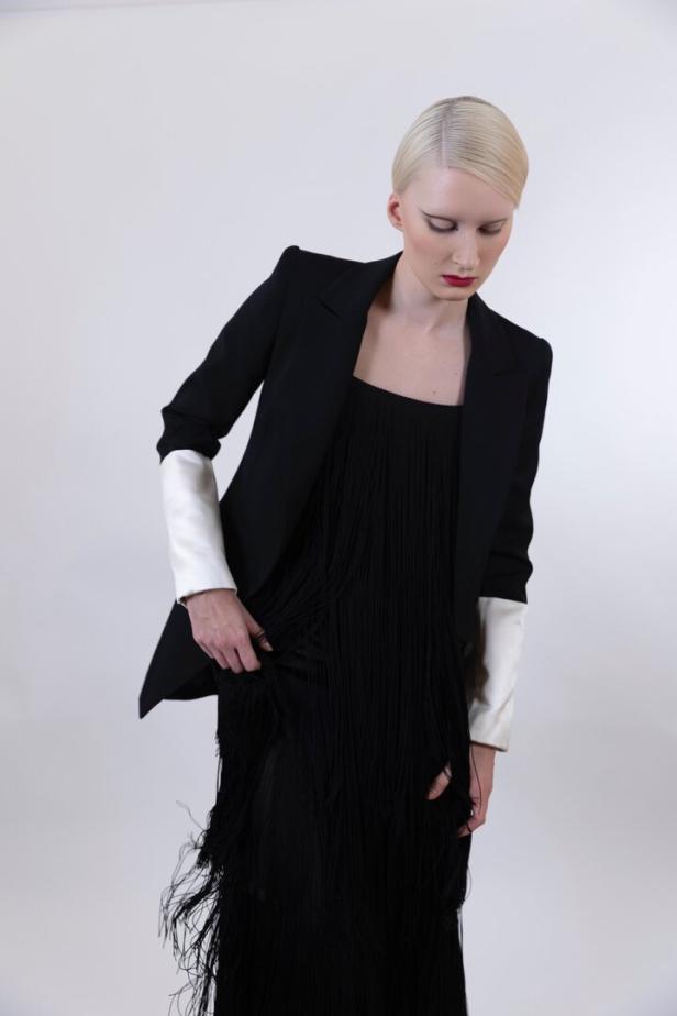 Eine Frau mit blonden Haaren trägt ein schwarzes Fransenkleid und einen Blazer mit weißen Ärmeln.