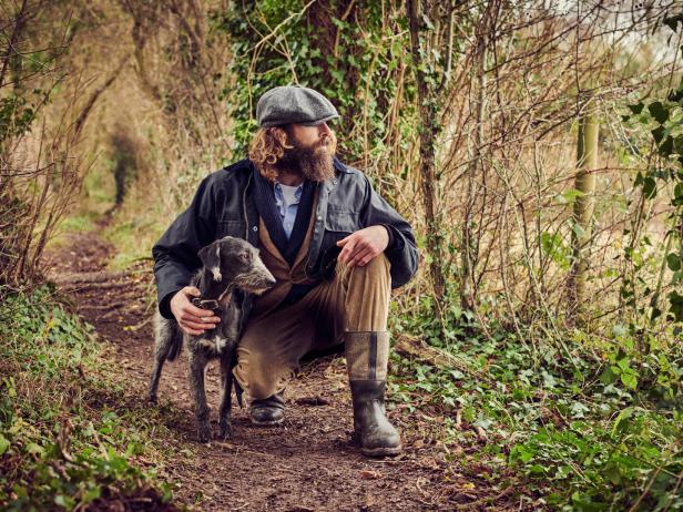Ein Mann mit Bart und Mütze kauert mit einem Hund auf einem Waldweg.