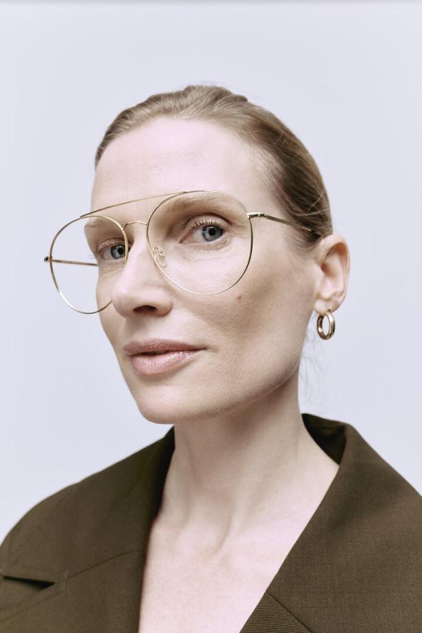 Eine Frau mit blonden Haaren und goldenen Ohrringen trägt eine goldene Brille und ein grünes Jackett.