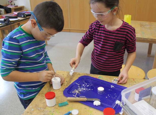 Zwei Kinder mit Schutzbrillen basteln an einem Tisch mit verschiedenen Materialien.