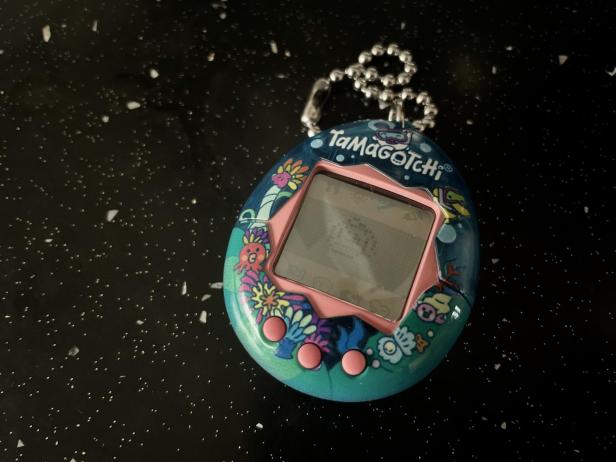 Ein blau-pinkes Tamagotchi mit Meeresmotiven liegt auf einer glitzernden Oberfläche.