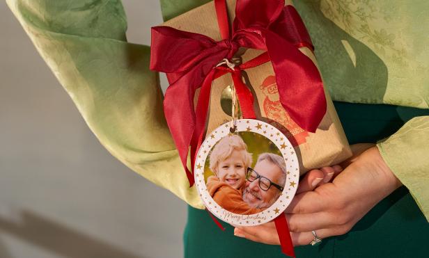 Eine Frau in einer grünen Bluse hält ein Weihnachtsgeschenk hinter ihrem Rücken versteckt. An diesem hängt ein Foto-Anhänger, auf dem Vater  und Sohn drauf sind.