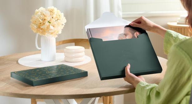 Eine Frau mit einer grünen Bluse sitzt an einem Tisch, auf dem ein Blumenstrauß steht. Sie packt gerade ein Weihnachtsgeschenk aus, in dem sich ein Fotobuch befindet.