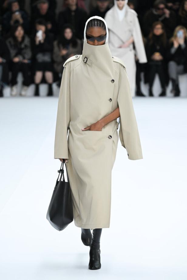 Eine Frau mit Sonnenbrille und einem beigen Trenchcoat mit hochgeschlossenem Kragen läuft auf einem Laufsteg.