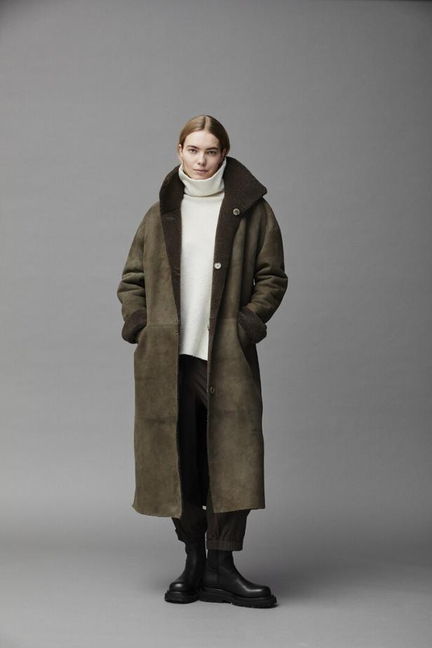 Eine Frau trägt einen langen, olivgrünen Shearling-Mantel über einem weißen Rollkragenpullover.