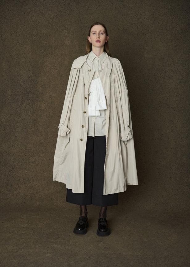 Eine Frau mit Trenchcoat, Hemd und Culottes posiert vor einem braunen Hintergrund.