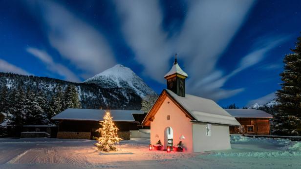 Eine verschneite Kapelle und ein beleuchteter Weihnachtsbaum vor einer Bergkulisse bei Nacht.