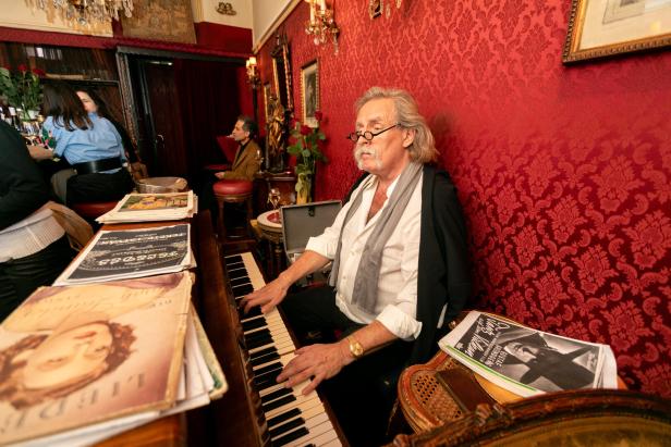 Ein älterer Mann mit Schnurrbart spielt Klavier in einem eleganten Raum.