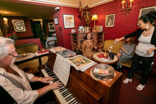 In einem gemütlichen Salon spielt ein Mann Klavier, während eine Frau Getränke serviert.