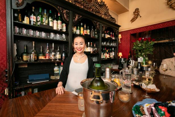 Eine lächelnde Frau steht hinter einer Bar mit Spirituosen und Gläsern.