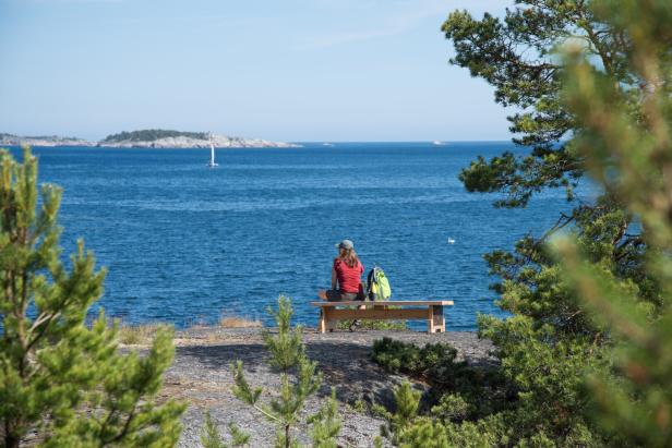 Frau sitzt auf einer Schwedischen Schäreninsel auf einer Bank mit Wanderrucksack und schaut aufs Meer