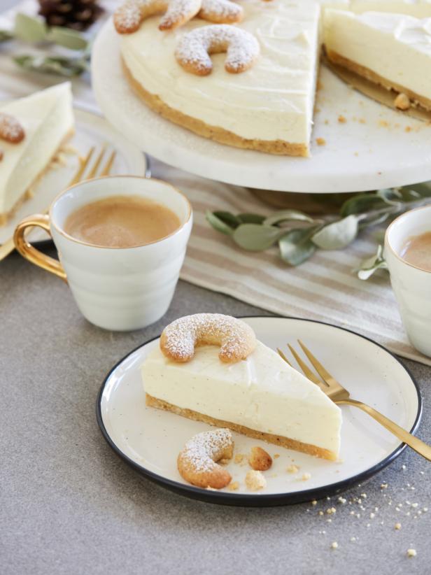 Ein Stück Käsekuchen mit Vanillekipferl auf einem Teller, begleitet von zwei Tassen Kaffee.