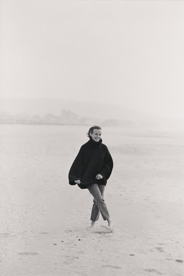 Eine Frau in einem übergroßen Pullover und Jeans geht barfuß am Strand entlang.