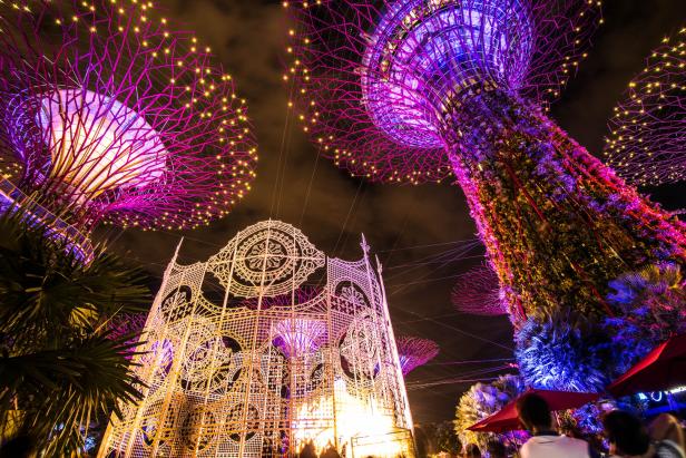 Beleuchtete künstliche Bäume und eine filigrane Lichtinstallation leuchten bunt in einer nächtlichen Parklandschaft in Singapur.