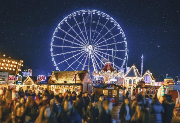 Ein belebter Weihnachtsmarkt mit einem leuchtenden Riesenrad im Hintergrund.