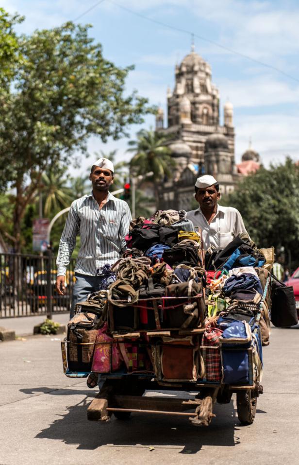 Zwei Männer schieben einen Karren voller Gepäck durch eine indische Stadt.
